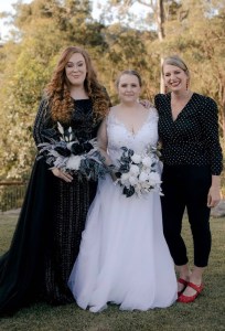 Roxy Celebrant wearing a black and white blouse marrying two brides, one wearing a white dress, one wearing a green